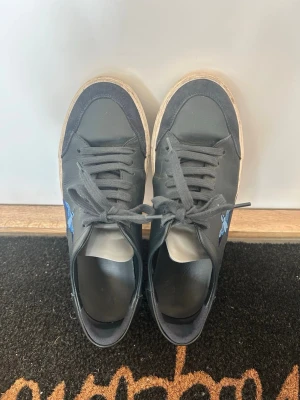 Axel Arigato svarta sneakers med fågel - Svarta sneakers från Axel Arigato med broderad blå fågel på sidan och texten 'ARIGATO'. Skorna har svarta skosnören, vit sula och detaljer i mocka framtill. Använda max 5 gånger