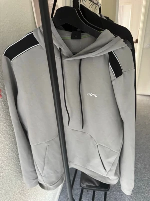 Grå hoodie från BOSS med svarta detaljer - Snygg grå hoodie från BOSS med svarta detaljer på axlarna och vita linjer. Klassisk huva med svarta dragsnören och en stor magficka framtill. BOSS-logga tryckt på bröstet. Perfekt för en avslappnad och trendig stil.