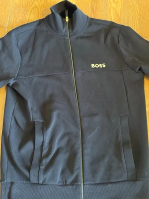 BOSS Hugo Boss Zip-up Sweatshirt / Track Jacket i svart med fin broderad guldlogga och matchande gulddragkedja. Storlek M, 100% bomull. - Stilren och klassisk zip-tröja med hög krage, ribbade muddar och praktiska fickor fram. Den har en skön och normal passform, mjukt material som andas bra och ett lyxigt utseende som passar perfekt till jeans, chinos eller som mellanlager. Plagget är i väldigt bra skick. Den är ren, fräsch, inga fläckar eller slitage och är nyligen tvättad. En tidlös BOSS-favorit som ser betydligt dyrare ut än priset. Pris: 529 kr