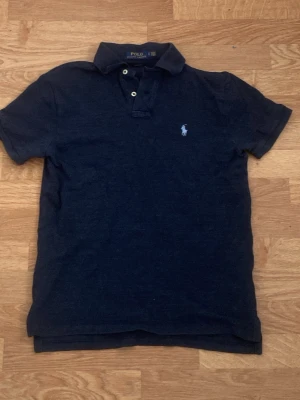 Mörkblå pikétröja från Ralph Lauren - Klassisk mörkblå pikétröja från Ralph Lauren med ljusblå broderad logga på bröstet. Tröjan har korta ärmar, krage och knappar framtill. Helt oandvänd utöver när jag provade på den för att se om den passa. Skickar inom 24 timmar