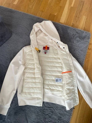 Vit pufferjacka med huva från Parajumpers - Snygg vit pufferjacka från Parajumpers med quiltad front, huva och dragkedja. Jackan har en sportig look med detaljer i orange och gul vid halsen. Perfekt för kyliga dagar och enkel att matcha till olika outfits.