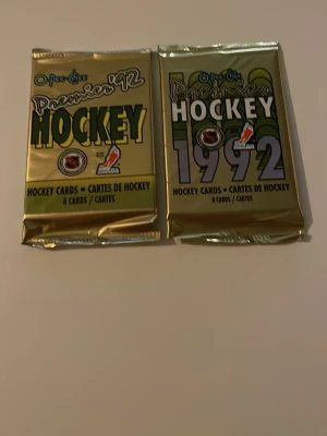O-Pee-Chee Premier 1992 Hockeykort - Två oöppnade paket O-Pee-Chee Premier 1992 hockeykort. Varje paket innehåller 8 samlarkort och är förpackade i guldfärgade folieomslag med tydlig grafik och text. Perfekt för dig som gillar samlarkort och hockeynostalgi.