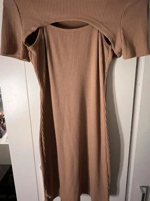 Beige ribbad klänning med cut out - Trendig beige ribbad klänning med kort ärm och cool cut out-detalj över bröstet. Klänningen har en kroppsnära passform och är tillverkad i ett stretchigt material som ger en snygg siluett. Perfekt för dig som vill sticka ut med en unik look.