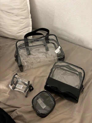 Set med transparenta väskor - Säljer ett set med fyra transparenta väskor i olika storlekar. Väskorna är gjorda i genomskinlig plast med svarta detaljer och handtag. Perfekt för att organisera småprylar eller använda som necessär. Snygg och modern design som passar till allt.