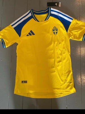 Sverige fotbollströja Adidas gul/blå - Gul fotbollströja från Adidas med blå och vita detaljer, officiella Sverige-emblemet på bröstet och Adidas-logga. Tillverkad i lätt och ventilerande material med Climalite-teknologi. Perfekt för match eller träning.