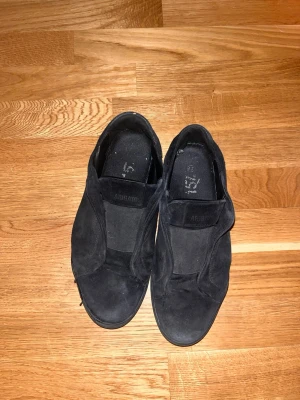 Svarta slip-on sneakers från ARIGATO - Stilrena svarta slip-on sneakers från ARIGATO i mocka med elastiska paneler på sidorna. Skorna har en låg siluett och en robust gummisula som ger en clean look. Perfekta för dig som gillar minimalistisk och modern stil.