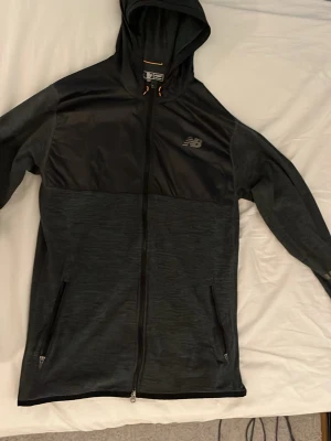 Svart luvtröja från New Balance - Snygg svart hoodie från New Balance med hel dragkedja och två fickor framtill. Materialet är en mix av slät och lätt glansig textil upptill och mjukare tyg nedtill. Klassisk huva och diskret logga på bröstet. Perfekt för en sportig och avslappnad stil.