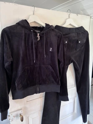 Svart velour hoodie och byxor från Juicy Couture - Mjuk och skön svart hoodie och mjukisbyxor i velour från Juicy Couture med dragkedja, huva med snören och fickor framtill. Ikonisk JC-logga broderad på bröstet. Byxorna har 2 fickor bak med knappar. Perfekt för en avslappnad och trendig look. Byxorna saknar snöre.