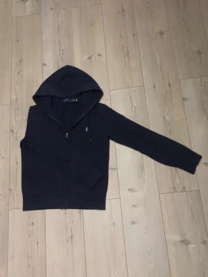 Mörkblå hoodie från Polo Ralph Lauren - Mörkblå hoodie med dragkedja från Polo Ralph Lauren. Klassisk design med huva och broderad logga på bröstet. Mjuk bomullskänsla och ribbade muddar vid ärmslut och midja. Perfekt för en avslappnad och stilren look.