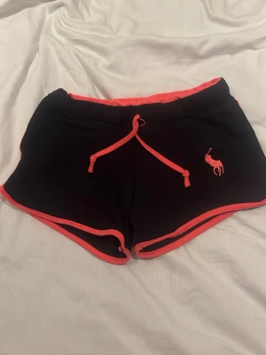 Svarta shorts från Ralph Lauren - Svarta shorts från Ralph Lauren med neonrosa detaljer och snörning i midjan. Klassisk broderad logga på ena benet och kontrasterande kantband. Perfekta för en sportig och avslappnad stil.