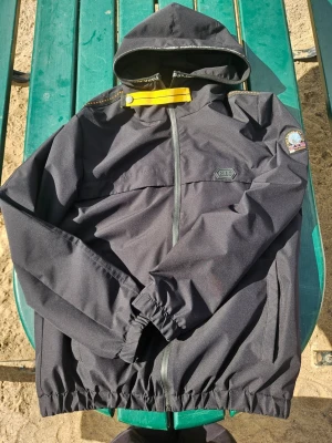 Svart vindjacka från Parajumper  - Svart vindjacka från Parajumper med huva, dragkedja och elastiska muddar. Jackan har en patch på ärmen och en diskret logga på bröstet. Materialet är lätt och vindtätt, perfekt för blåsiga dagar. Snygga detaljer som gul rem vid kragen. Strl. M