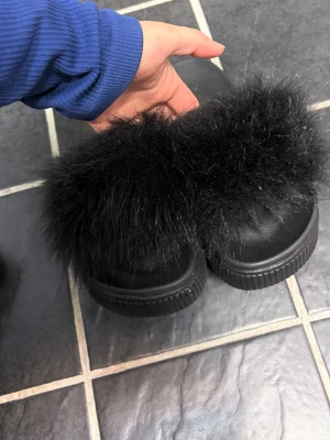 Svarta fluffiga tofflor från Vox - Säljer ett par svarta tofflor från Vox med bred rem i mjukt, fluffigt fuskpälsmaterial. Sulan är i svart gummi och tofflorna har en modern slip-in design. Perfekta för att chilla hemma eller när du vill ha något extra comfy på fötterna.