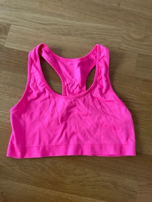Neonrosa sport-bh med brottarrygg - Snygg neonrosa sport-bh med brottarrygg och bred resår nedtill. Perfekt för träning och aktiviteter där du vill ha bra stöd och rörelsefrihet. Tillverkad i stretchigt material som andas och sitter skönt mot kroppen.