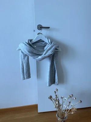 Ljusgrå mjuk halsduk - En enkel och stilren ljusgrå halsduk i mjukt material. Halsduken är lång och rektangulär, perfekt att vira flera varv runt halsen. Passar till de flesta outfits och ger en clean look. Märke och exakt material är okänt men tyget ser ut att vara stickat och skönt.