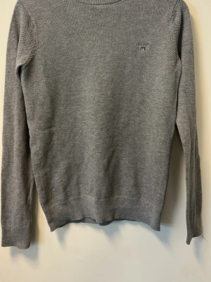 Grå stickad tröja från Gant - Snygg grå stickad tröja från Gant med diskret logga på bröstet. Tröjan har rund halsringning, långa ärmar och ribbade muddar. Perfekt till jeans eller kjol för en clean look.