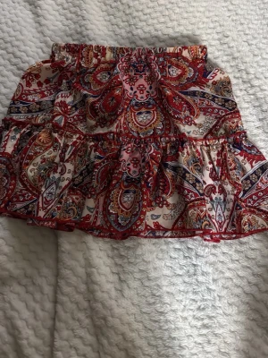 Mönstrad volangkjol  - Kort kjol med volang och elastisk midja. Färgglatt paisleymönster i rött, beige, blått och inslag av rosa. Lätt och luftig känsla, perfekt för varma dagar. Kjolen har en feminin och ungdomlig stil med fina detaljer.