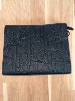 Dior Clutch Oblique Black - Säljer en exklusiv Dior clutch i svart präglat läder med ikoniskt Oblique-mönster. Stilren och tidlös modell som passar både vardag och mer uppklädda tillfällen.  Färg: Svart Material: Läder Skick: Mycket fint skick, sparsamt använd. Inga tydliga skador. Dragkedja fungerar perfekt. Invändigt med kortfack och bra förvaring.  Perfekt som clutch, necessär eller för att organisera i större väska.