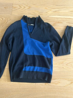 Halv zip från J.Lindeberg - Snygg marin blå tröja från J.Lindeberg med blåa grafiska detaljer framtill och på ryggen. Tröjan har halv dragkedja vid halsen, ribbade muddar och en modern, sportig look. Perfekt för dig som gillar stilrena men ändå unika plagg.