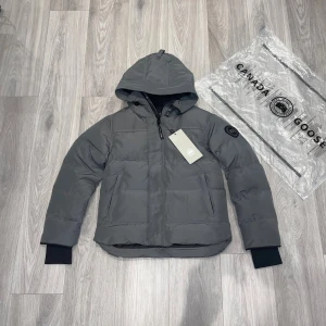 Grå pufferjacka från Canada Goose - Grå pufferjacka från Canada Goose med huva och svart patch på ärmen. Jackan har quiltad design, dragkedja framtill och ribbade muddar vid ärmslut. Perfekt för kalla dagar och har en clean, modern look. Pris kan diskuteras vid snabb affär