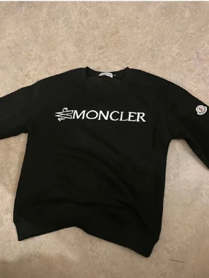 Svart Moncler sweatshirt - Snygg svart sweatshirt från Moncler med stor vit logga framtill och Moncler-märke på ena ärmen. Klassisk rund halsringning och ribbade muddar vid ärmslut och nederkant. Perfekt för en trendig och avslappnad stil.