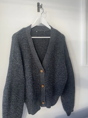 Mörkgrå stickad kofta med knappar - Mysig mörkgrå stickad kofta med v-ringning och stora bruna knappar framtill. Koftan har en avslappnad passform, ribbade muddar och är perfekt att slänga över en t-shirt. Passar till jeans eller kjol för en chill look. I storlek M men passar xs-m