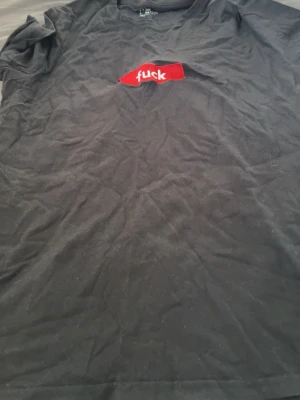 Svart t-shirt med röd brodyr - Svart t-shirt med ett broderat rött märke på bröstet där det står 'fuck' i vit text. Klassisk passform och rund halsringning. Perfekt för dig som vill ha en statement-tee med attityd.