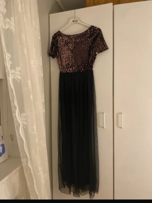 Svart långklänning med paljetter - Snygg svart långklänning med korta ärmar och glittrig ovandel täckt av guldiga paljetter. Kjoldelen är i tunn, genomskinlig polyester som ger ett elegant fall. Perfekt för dig som vill sticka ut med en glamorös look. 