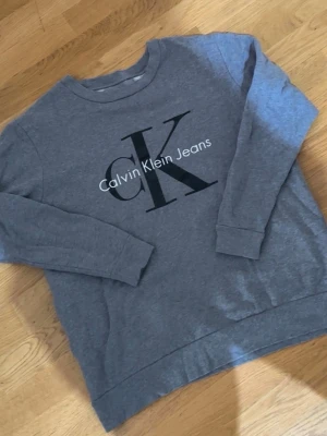 Grå sweatshirt från Calvin Klein Jeans - Säljer en grå sweatshirt från Calvin Klein Jeans med stort CK-tryck på bröstet. Tröjan har rund halsringning, långa ärmar och ribbade muddar vid ärmslut och nederkant. Perfekt för en avslappnad och stilren look.