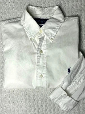 Ralph Lauren vit skjorta – Custom Fit (S) - Klassisk vit skjorta från Ralph Lauren i modellen Custom Fit, storlek S. Button-down krage och mörkblå broderad logga på bröstet. Stilren och lätt att matcha, funkar både uppklätt och vardagligt.  Skick: Bra begagnat skick, normalt bruksslitage Material: 100% bomull                        Bröstmått - ca 53 cm Längd - ca 73 cm Ärmlängd - ca 61 cm