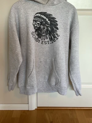 Grå hoodie med tryck och huva - Säljer en grå hoodie från SCNDI med stor svart tryck framtill som föreställer ett huvudbonadsmotiv och texten 'SCNDI EST. 2024'. Hoodien har klassisk känguruficka, ribbade muddar och en skön huva. Perfekt för dig som gillar streetwear och vill ha en avslappnad stil.