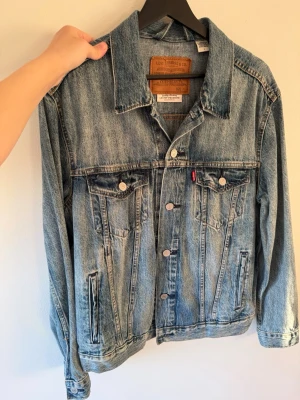 Levi's klassisk blå jeansjacka - Säljer en klassisk blå jeansjacka från Levi's med raka sömmar, två bröstfickor med knappar och två sidofickor. Jackan har silverfärgade knappar med logga och den ikoniska röda Levi's-taggen på bröstet. Perfekt oversized passform och tidlös look.