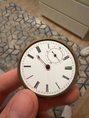 Antik fickur med romerska siffror - Säljer ett klassiskt antikt fickur med vit urtavla och svarta romerska siffror. Uret har en guldfärgad baksida med graverad detalj och en mindre sekundvisare. Perfekt för dig som gillar vintage och tidlös stil.