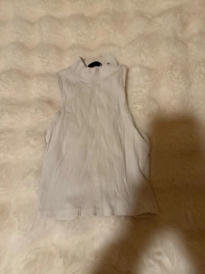 Vit ribbad ärmlös polotopp Zara - Säljer en vit ribbad polotopp från Lager 157 i storlek XS/S. Toppen är ärmlös med hög hals och har en tight passform, magtröja. Perfekt att styla med jeans eller kjol för en clean och trendig look.