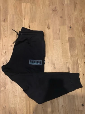 Svarta mjukisbyxor från Hoodrich - Säljer ett par svarta mjukisbyxor från Hoodrich med dragsko i midjan och ribbade muddar vid bensluten. Byxorna har en stor logga på vänster lår och är tillverkade i mjukt material som passar perfekt för chill dagar. Snygg och avslappnad passform.
