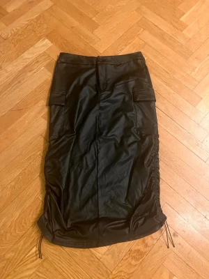 Svart långkjol i skinnimitation - Säljer en svart långkjol i skinnimitation med coola snörningsdetaljer i sidorna och fickor framtill. Kjolen har en rak passform och ger en edgy vibe till din outfit. Perfekt för dig som vill sticka ut lite extra.Skriv gärna för mått! 💕