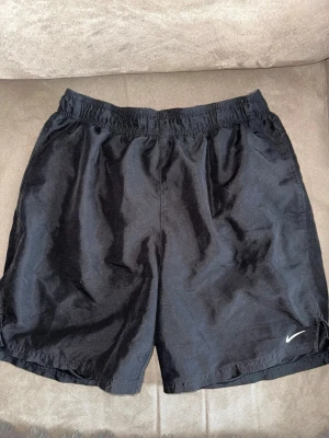 Svarta shorts från Nike - Säljer ett par klassiska svarta shorts från Nike med elastisk midja och liten vit logga på benet. Perfekta för träning eller chill dagar. Tillverkade i lätt och bekvämt syntetmaterial som andas.