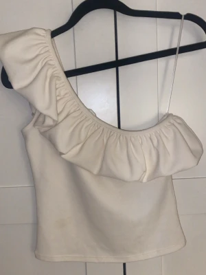Vit oneshoulder topp från H&M - Säljer en vit oneshoulder topp från H&M i storlek XS. Toppen har en bred volang längs axeln och ett smalt axelband på ena sidan. Perfekt för dig som vill ha en unik och trendig look. Materialet känns mjukt och bekvämt. - Tröjan har en liten fläck på framsidan 