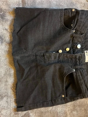  jeanskjol - Säljer en svart kort jeanskjol från gina tricot. Kjolen har rå kant nertill, knappgylf med silverfärgade knappar och klassiska jeansfickor fram. 