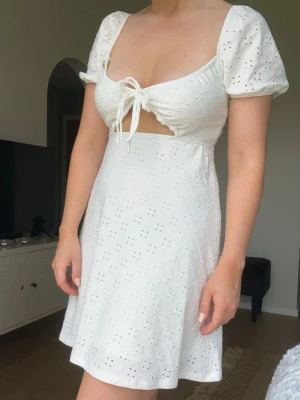 Vit kort klänning med puffärm från H&M, stl S - Söt vit kort klänning i bomull med broderade hål, puffärmar och knyt framtill. Klänningen har en lätt utsvängd kjol och öppen detalj under bysten. Sitter superbra runt bysten och ramlar inte av! 92% polyester och 8% elastan. 🩷