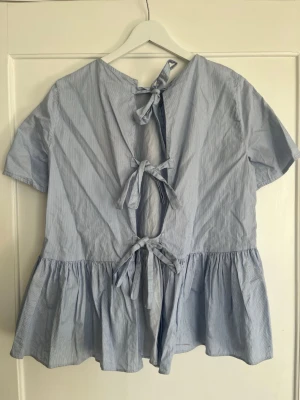 Ljusblå volangblus med knytband - Supersöt ljusblå blus med tunna vita ränder och volang nedtill. Blusen har korta ärmar och tre dekorativa knytband i ryggen som ger en unik look. Tillverkad i ett lätt bomullstyg som känns luftigt och skönt. Perfekt för dig som gillar en avslappnad men trendig stil.