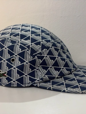 Lacoste keps - Snygg keps i blått och vitt med geometriskt mönster och klassisk Lacoste-logga på sidan. Kepsen har böjd skärm och metallöljetter för ventilation. Perfekt accessoar för en trendig streetwear-look.