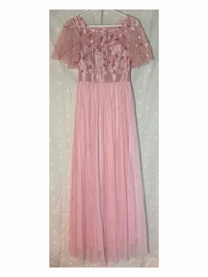 Lång rosa klänning - Romantisk långklänning i ljusrosa med transparenta, broderade ärmar och glittriga detaljer. Klänningen har ett vackert blommigt mönster upptill, markerad midja med satinband och ett svepande, tunt tyg i kjolen. Perfekt för speciella tillfällen!