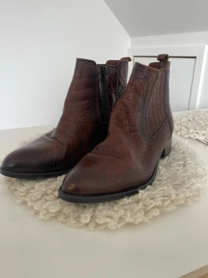 Bruna cowboy boots i ormmönstrat läder - Säljer ett par bruna cowboy boots i ormmönstrat läder med spetsig tå och låg klack. Bootsens yta har en snygg struktur och dragkedja på sidan för enkel på- och avtagning. Perfekta för dig som vill ha en cool westernstil.