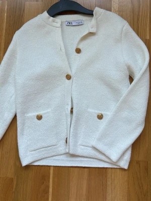 Vit kofta med guldknappar från Zara - Stilren vit kofta från Zara med fyra dekorativa guldknappar framtill och två fickor. Koftan har rund halsringning och är tillverkad i ett mjukt, strukturerat material. Perfekt att slänga över en topp för en clean look.