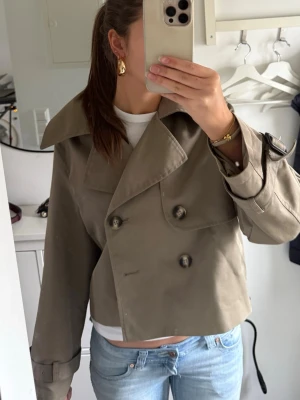 Kort grön trenchcoat - Snygg kort trenchcoat i olivgrön med dubbelknäppning. Jackan har justerbara remmar vid ärmsluten och en modern passform. Perfekt att slänga över en t-shirt för en cool och avslappnad look. Materialet är slätt och klassiskt trenchcoat-tyg.