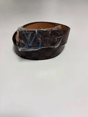 Brunt rutigt IV bälte med metallspänne - Snyggt brunt bälte från Iouis Vuitton med klassiskt rutigt mönster och stort IV-spänne i metall. Bältet har en exklusiv känsla och passar perfekt till jeans eller kostymbyxor. Insidan är ljusbrun och materialet känns lyxigt.