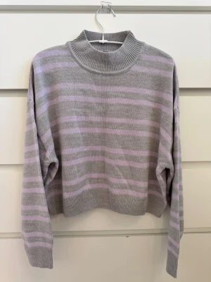 Grå och lila randig stickad tröja - H&M - Snygg stickad tröja i grått med lila horisontella ränder. Tröjan har en hög, ribbad krage och långa ärmar med ribbade muddar. Passformen är lite boxig och avslappnad, perfekt för en chill look. Materialet känns mjukt och bekvämt 💜🩶