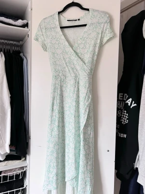 Mintgrön blommig omlottklänning - Säljer en söt mintgrön omlottklänning med blommigt mönster. Klänningen har knytband i midjan för en perfekt passform. Finns en liten fläck bak på ryggen (se sista bilden). Oanvänd!
