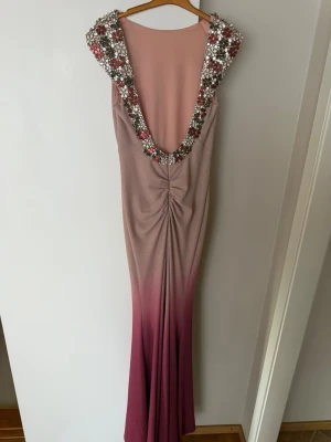 Lång rosa klänning med pärldetaljer - Säljer en elegant långklänning i rosa och lila/rosa toner med djup öppen rygg. Klänningen har pärl- och paljettdekorationer runt axlar och rygg, samt en snygg ombre-effekt från ljusrosa till lila/rosa. Perfekt för bal eller festliga tillfällen. Aldrig använd!!! Köpt i Bosnien