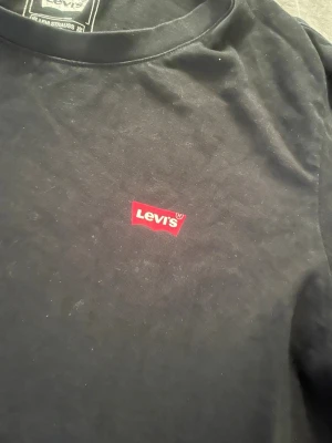 Svart Levi's t-shirt med logga - Klassisk svart t-shirt från Levi's med den ikoniska röda loggan på bröstet. T-shirten har rund halsringning och är tillverkad i mjuk bomull för en skön känsla. Perfekt basplagg för alla tillfällen och den är storlek S 